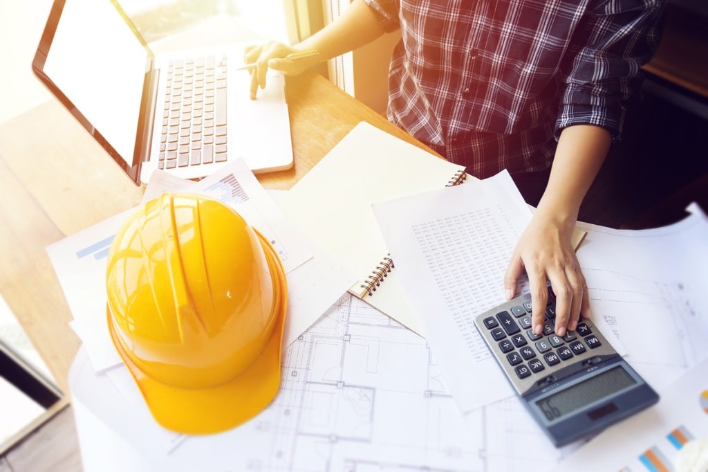 Asian engineer , architect woman using calculator for accounting and analyzing building structure from blueprint and paper chart in front of computer laptop at office work space.